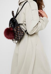 Sac à dos noir et marron à motif avec un pompon en fourrure, équipé de bretelles réglables et d'une fermeture éclair. Tissu texturé et détails métalliques.