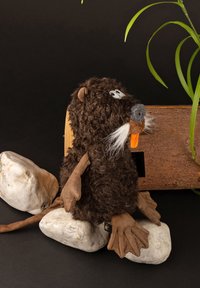 Plush beaver toy, brown fur, gray snout, orange nose, white whiskers, brown leather-like paws, seated on white stones with wood background.