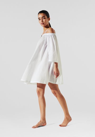 Robe blanche décolletée avec des manches bouffantes longues, fabriquée en tissu léger, se distingue par sa coupe ample et sa silhouette fluide.