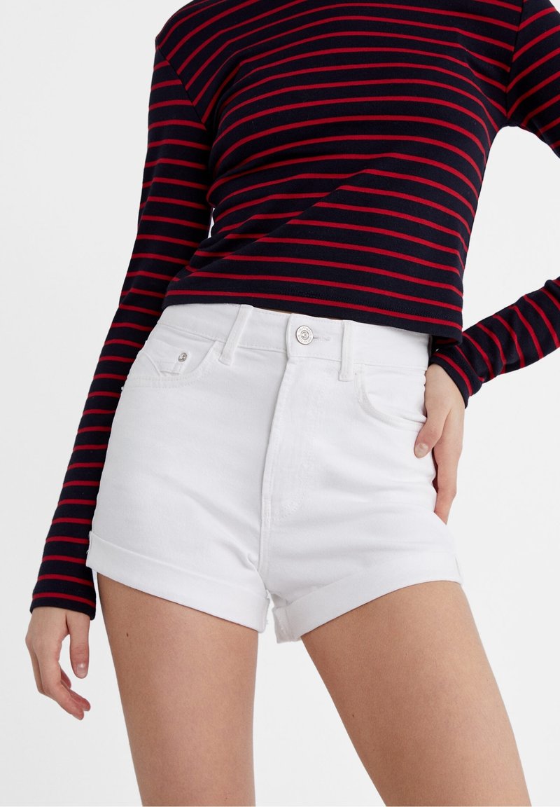 Femme portant un haut bleu marine à manches longues avec des rayures rouges et un short en denim blanc taille haute aux ourlets retroussés, debout avec la main sur la hanche.