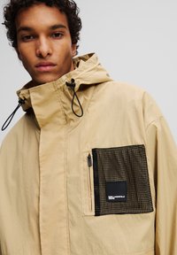 Beige Kapuzenjacke mit einer strukturierten schwarzen Mesh-Tasche und einem kleinen Logo-Patch. Verfügt über verstellbare Kordeln und eine glatte Stofftextur.