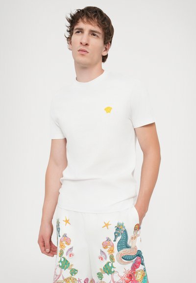White cotton t-shirt with a round neck and short sleeves, featuring a small yellow emblem on the chest. Textured, lightweight fabric.