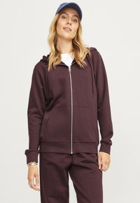 Felpa con zip bordeaux dotata di cappuccio regolabile, tasche anteriori e una texture morbida, abbinata a pantaloni coordinati e un cappellino blu.