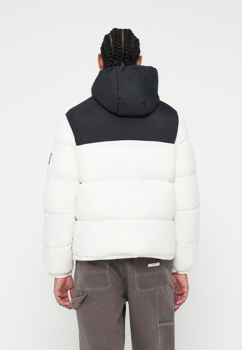 Biała kurtka puchowa z czarną górną częścią, z kapturem, pikowanym wzorem i elastycznymi mankietami. Nosi się z szarymi spodniami cargo.