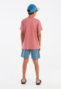 Rode heide T-shirt, losse pasvorm, gecombineerd met blauwe katoenen shorts. Kind draagt een lichtblauw en beige pet met zichtbare stiksels en hardware.