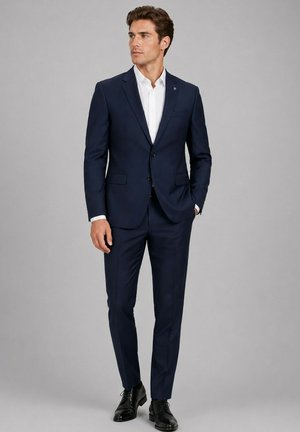 Homme portant un costume bleu foncé ajusté avec une chemise blanche et des chaussures noires, debout avec une main dans sa poche.