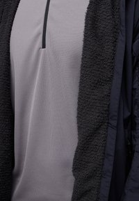 Ein graues, leichtes Oberteil mit einem zentralen schwarzen Reißverschluss und einer dunklen, strukturierten Außenlage. Die Textur steht im Kontrast zum glatten Stoff darunter.