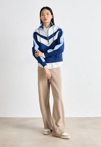 Giacca zip bianca e blu con design a zigzag, abbinata a pantaloni larghi beige e sneakers bianche, caratterizzata da uno stile pulito e casual.