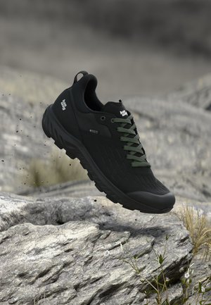 Juodi žygio batai su žaliais raišteliais ore virš uolėto paviršiaus, su Gore-Tex ženklu, užtikrinančiu nepralaidumą vandeniui ir orui laidumą.