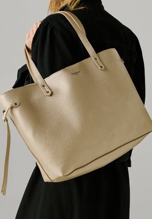 Mujer con chaqueta negra lleva un gran bolso tote de cuero beige texturizado con asas dobles sobre el hombro.