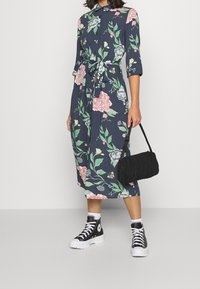 Blommönstrad klänning med trekvartsärmar, marinblå bakgrund, bälte i midjan, tillsammans med svart väska och högskurna sneakers.