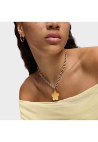 Collana in argento con un pendente a forma di fiore giallo. Indossata sopra una morbida maglietta gialla off-shoulder. Design semplice con una finitura lucida.