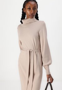 Abito beige lavorato a maglia con colletto alto, vita cintata e maniche lunghe leggermente a sbuffo. Tessuto morbido, design minimalista, lunghezza a metà polpaccio.