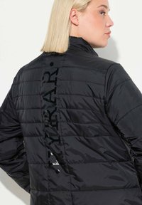 Sort pufferjakke med vandret quiltede stikninger, høj krave og et stort vertikalt logo langs ryggen i en blank finish.