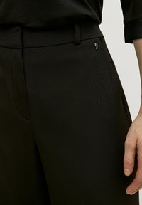 Czarne, dopasowane spodnie o gładkiej fakturze i subtelnych pionowych prążkach, z małym metalowym logo przy pasku.