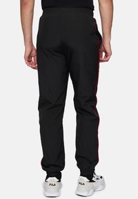 Zwarte sportbroek met een gladde textuur, voorzien van rode zijstrepen, een elastische tailleband en elastische boorden. Draagt een paar witte sneakers.
