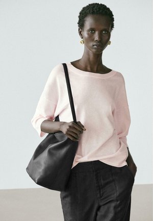 Frau mit kurzen lockigen Haaren trägt hellrosa Pullover, schwarze Hose, goldene Ohrringe und hat eine große schwarze Umhängetasche dabei.