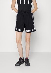 Pantalones cortos deportivos negros con franjas laterales blancas, cinturilla elástica y cordón. Combinados con zapatos grises y negros. Diseño ajustado.