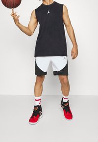 Haut sans manches noir, shorts blanc et noir, chaussures de basketball rouge et noir, et chaussettes blanches avec des accents rouges. Tenant un ballon de basketball.