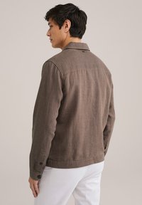 Brown textured jacket with a collar, button cuffs, and a straight fit. Features a subtle herringbone pattern and a tag at the back.