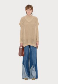 Beżowy sweter wykonany na drutach z szerokim dekoltem, w połączeniu z niebieskimi spodniami typu wide-leg w fasonie z kolorowym blokiem. Dodatki obejmują brązową torebkę.