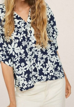 Kvinde iført en marineblå bluse med hvidt blomsterprint og cremefarvede bukser, stående foran en neutral baggrund.