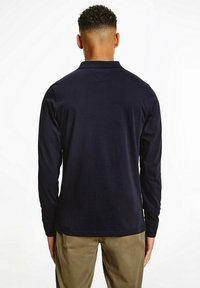 Marineblaues Langarm-Polohemd mit weicher Textur, klassischem Kragen und seitlichen Schlitzen am Saum. Die Rückansicht zeigt minimale Designdetails.