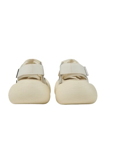 Paire de chaussures couleur crème avec des bouts arrondis et de larges brides sur le dessus, photographiées de face sur un fond blanc.