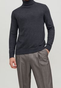Dunkelgrauer Rollkragenpullover in einer gerippten Stricktextur, kombiniert mit hellgrauen gestreiften Hosen. Die Hose hat Falten und Seitentaschen.