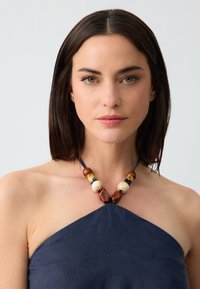 Collana di perline con perle in legno naturale, crema e marrone. Indossata con un top all'americana blu navy con un tessuto texture. Design semplice ed elegante.