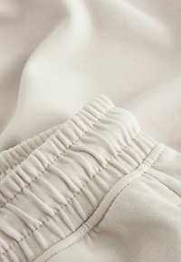 Beige elastisk linning med rynket tekstur, blødt bomuldsstof der falder i bløde folder, diskrete syningdetaljer synlige.