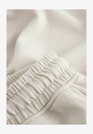 Bež elastični pas z gubano teksturo, mehak bombažni material, ki se nežno zloži v mehke gube, vidni subtilni detajli šivov.