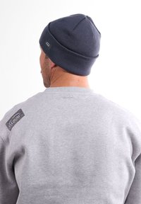 Sweat à capuche gris avec un patch logo sur l'épaule gauche et un bonnet en tricot bleu marine. Le bonnet a un sommet arrondi et une texture côtelée.