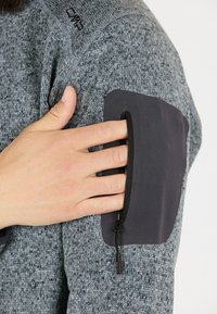 Grå tekstureret stof sweatshirt med en mørkegrå lynlås lomme på overarmen. Subtil syning detaljer på skulderen.