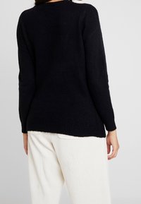 Persona che indossa un maglione nero a maniche lunghe e pantaloni bianchi a coste, vista di schiena su uno sfondo leggero e semplice.