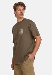Brown cotton T-shirt with a crew neck and short sleeves, featuring a graphic print in yellow on the left chest area. Casual fit.