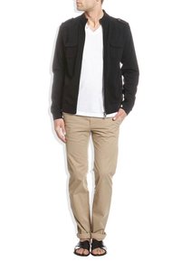 Svart zip-up jacka med två framfickor, bärs över en vit t-shirt, kombinerad med khakibyxor och svarta sandaler. Modellen står vänd framåt.