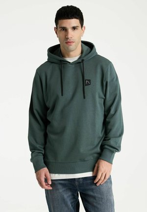 Jeune homme aux cheveux noirs courts portant un sweat à capuche vert et un jean bleu, debout devant un fond blanc.