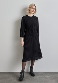 Schwarzes Faltenkleid mit langen Ärmeln, durchgehender Knopfleiste, tailliertem Bund und glatter Textur. Kombiniert mit schwarzen Knöchelstiefeln.