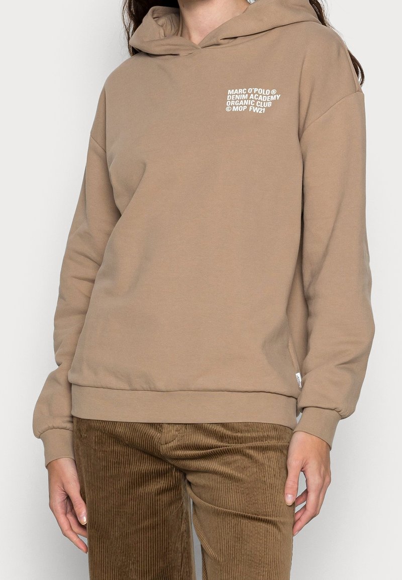 Personne portant un sweat à capuche beige avec un texte blanc sur la poitrine et un pantalon en velours côtelé marron, debout les bras détendus le long du corps.