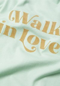 Tissu vert clair avec un texte doré indiquant "Walk in Love" dans une police à empattement décorative, présenté avec des plis doux.