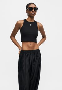 Γυναίκα που φοράει μαύρο αμάνικο crop top και φαρδιά μαύρα παντελόνια με γυαλιά ηλίου, στέκεται με τα χέρια πίσω από την πλάτη σε λευκό φόντο.