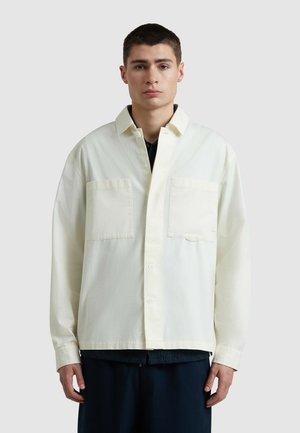 Camicione oversize color crema chiaro con due tasche sul petto, chiusura a bottoni e maniche lunghe. Realizzato in tessuto morbido con una leggera texture.