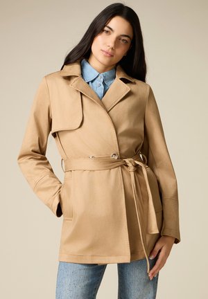 Frau trägt einen beigen Trenchcoat mit Gürtel über einem hellblauen Hemd mit Knöpfen und verzierter blauer Jeans, steht vor einem schlichten Hintergrund.