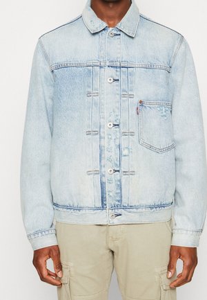 Man draagt lichtblauw spijkerjack met knopen aan de voorkant en een borstzak, gecombineerd met beige cargobroek.