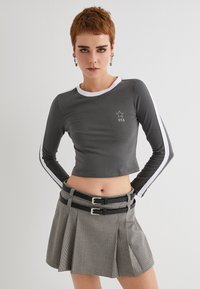 Grå langærmet crop top med hvide accenter og USA-logo; parret med en plisseret houndstooth nederdel med dobbelte spænder i sort.