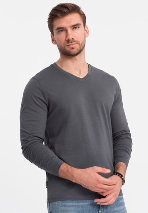 Camisa gris de manga larga con cuello en V, hecha de tela suave, con mangas ajustadas y un diseño limpio, combinada con jeans de mezclilla y una pulsera de cuentas.