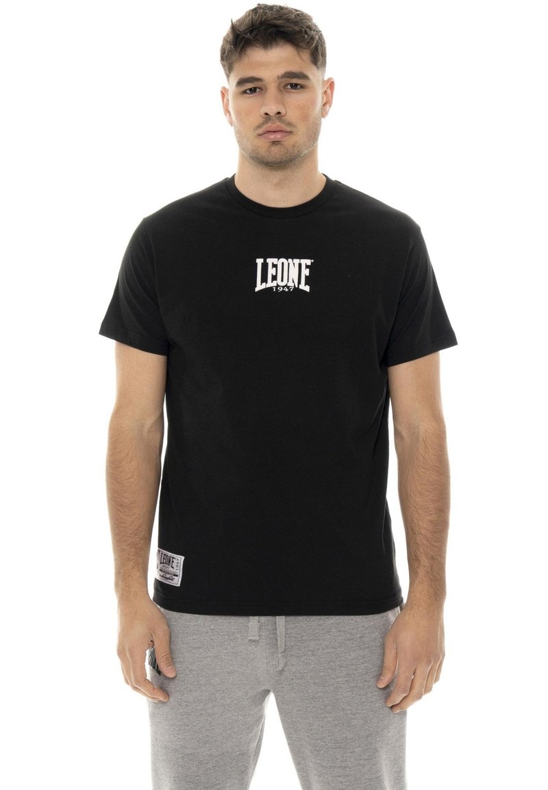 Un homme présente un T-shirt noir Leone 1947 et un pantalon de jogging gris sur fond blanc.