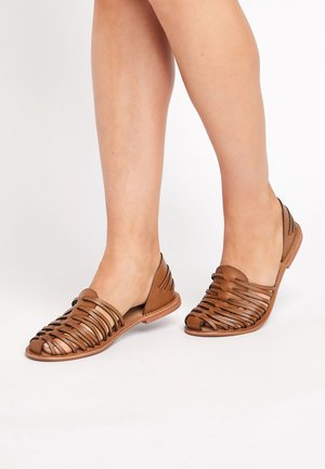 Zapatos de cuero marrón tipo slip-on con puntera abierta y diseño con recortes. Presentan una parte superior texturizada y suela plana. Ideales para clima cálido.