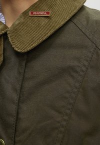 Veste vert olive avec un col en velours côtelé et une épingle "BEADNELL" rouge et or sur le col.